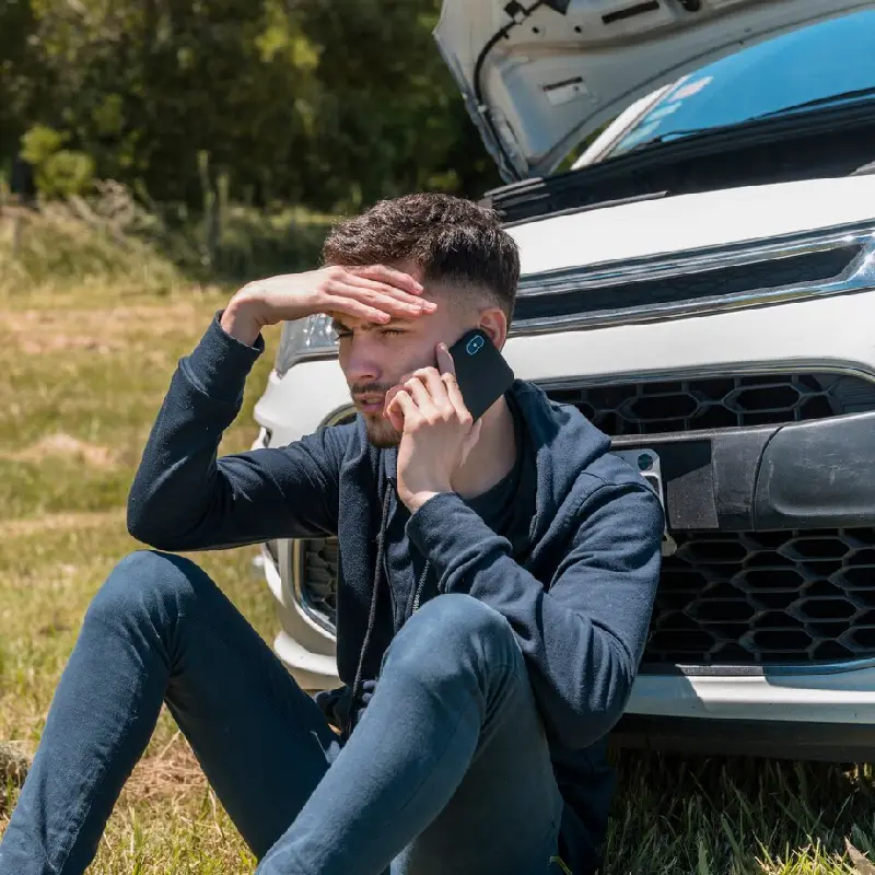 Man sitting beside a white car with the hood raised, looking stressed while talking on a smartphone, symbolizing the aftermath of a car accident and the need for legal assistance.