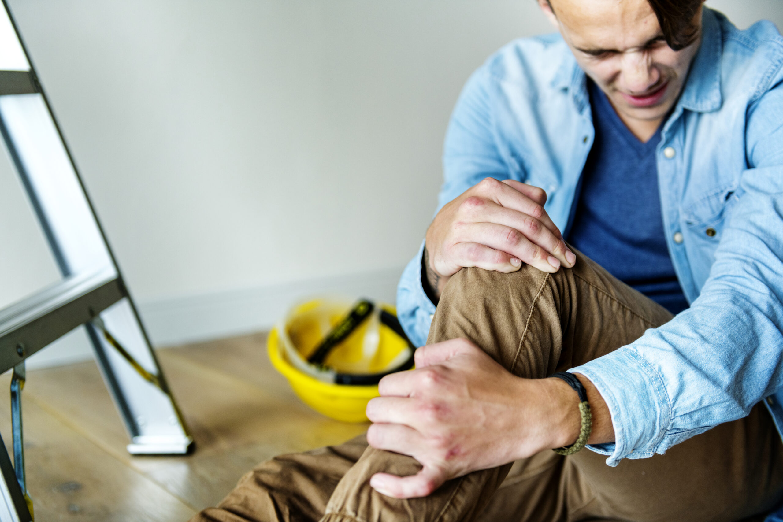 Man sitting on the floor, holding his knee in pain, with a ladder and a yellow hard hat in the background, illustrating personal injury in a home safety context.