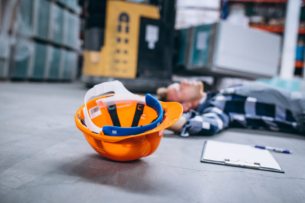 Orange safety helmet on warehouse floor with injured man lying nearby, illustrating workplace accident for personal injury legal services in Munger Place.