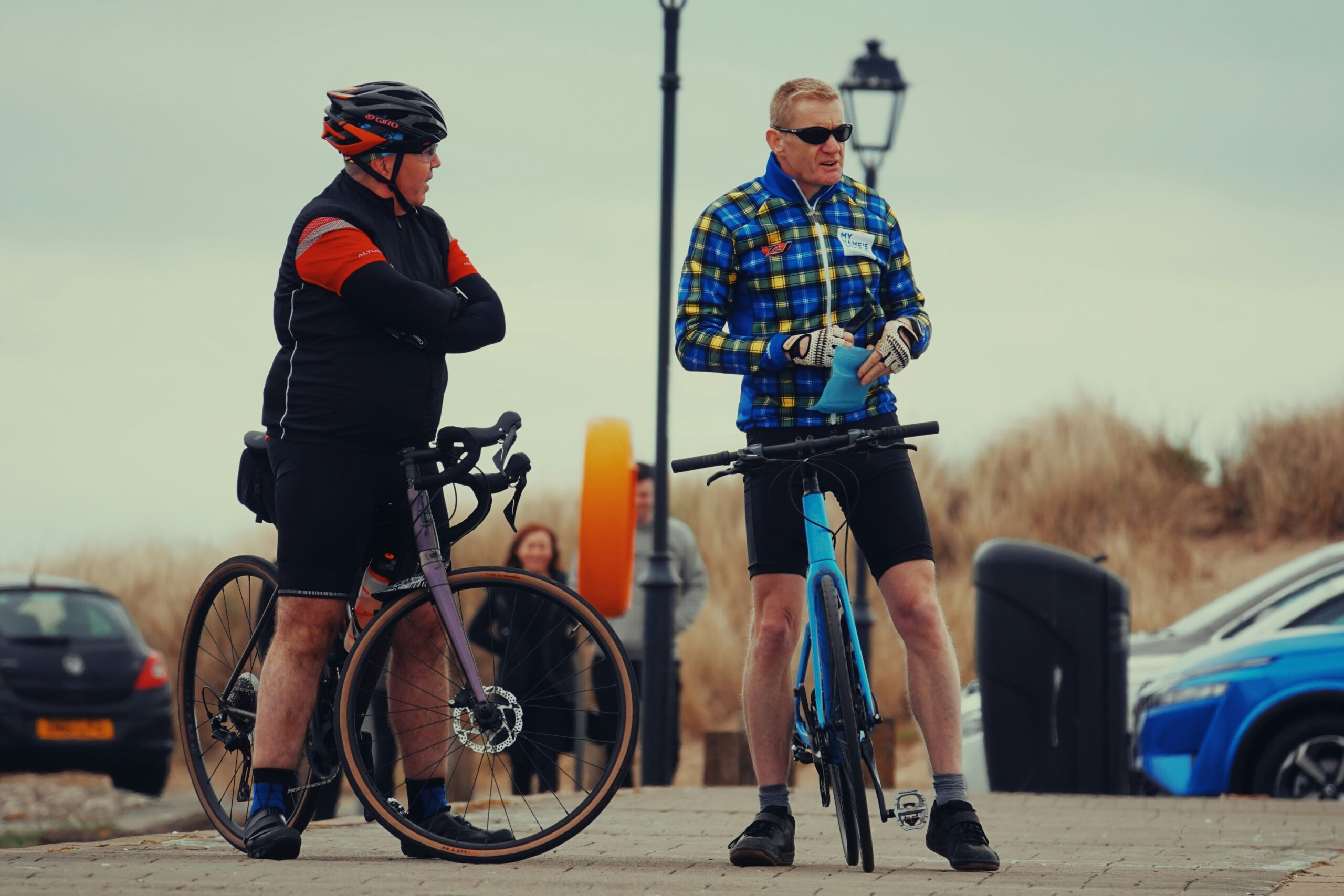 Two cyclists conversing, one in a black and red outfit with arms crossed, the other in a blue and yellow plaid jacket, both near bicycles, highlighting the importance of bicycle safety in Pearland's busy roads.