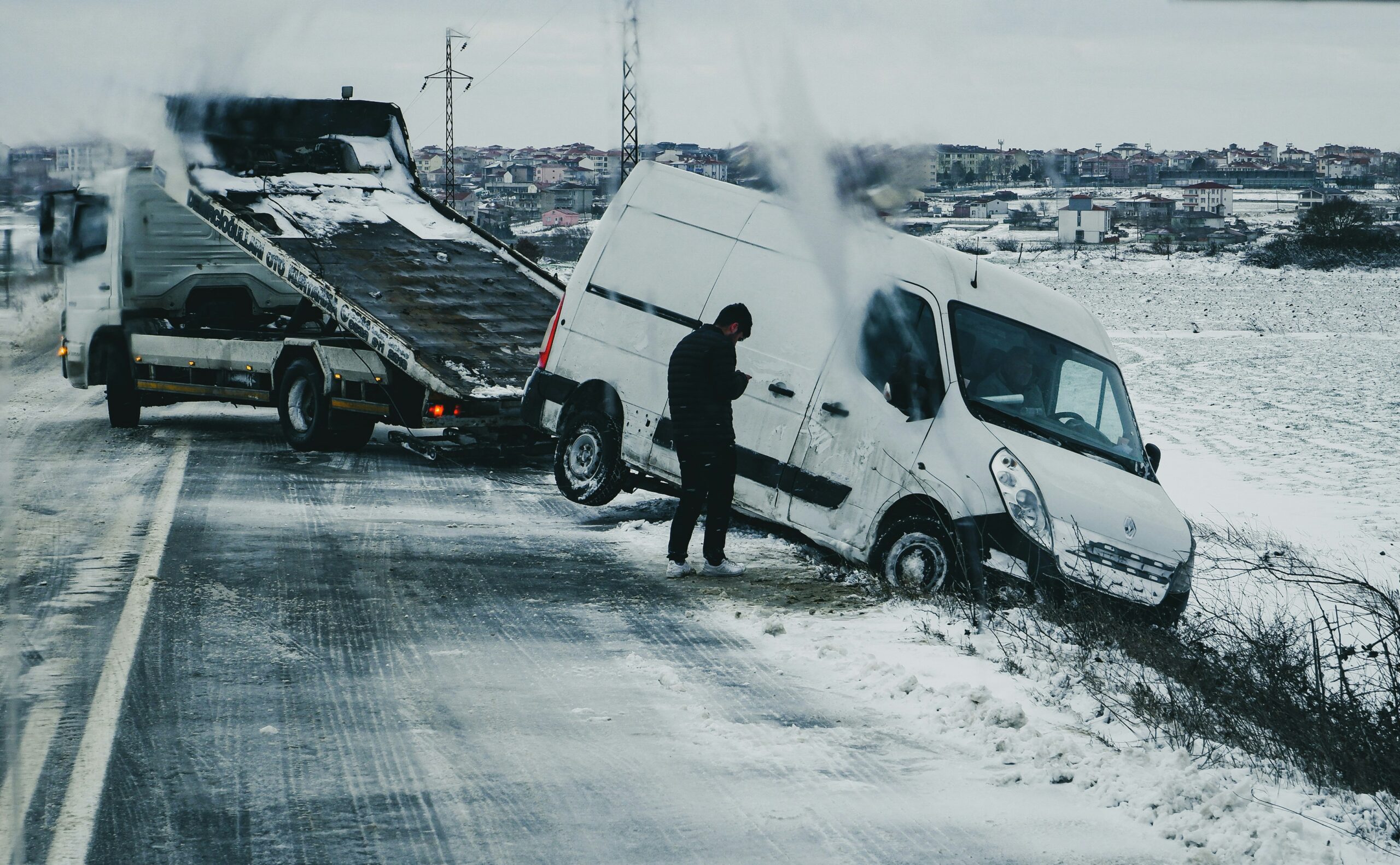 Snowy roadside accident scene with a white van partially in a ditch and a tow truck nearby, illustrating vehicle recovery efforts in adverse weather conditions.
