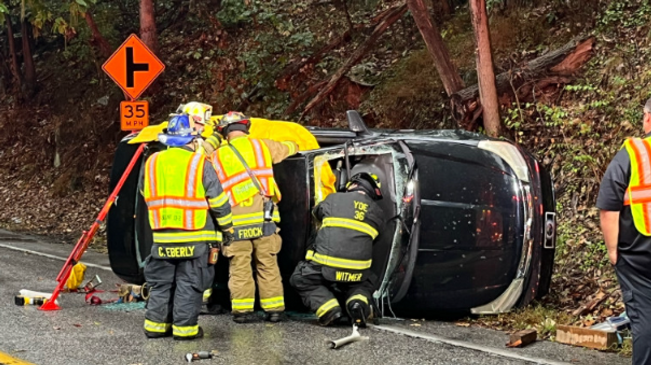 Emergency responders assisting at a rollover accident scene with a black vehicle on its side, surrounded by safety equipment and a warning sign indicating a sharp turn ahead.