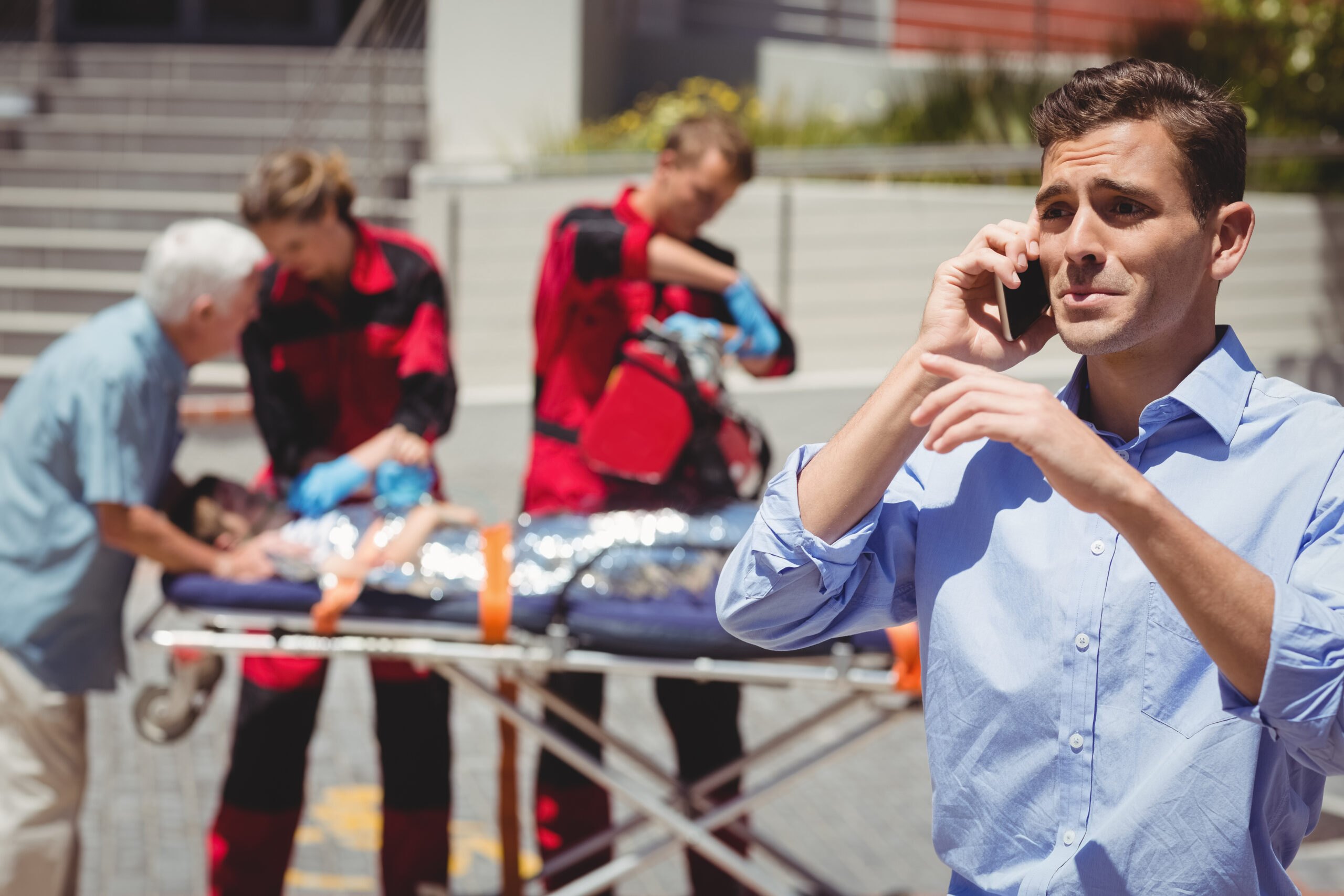 Man talking on mobile phone in foreground, paramedics attending to injured boy on stretcher in background, emergency scene in Westmoreland Heights.
