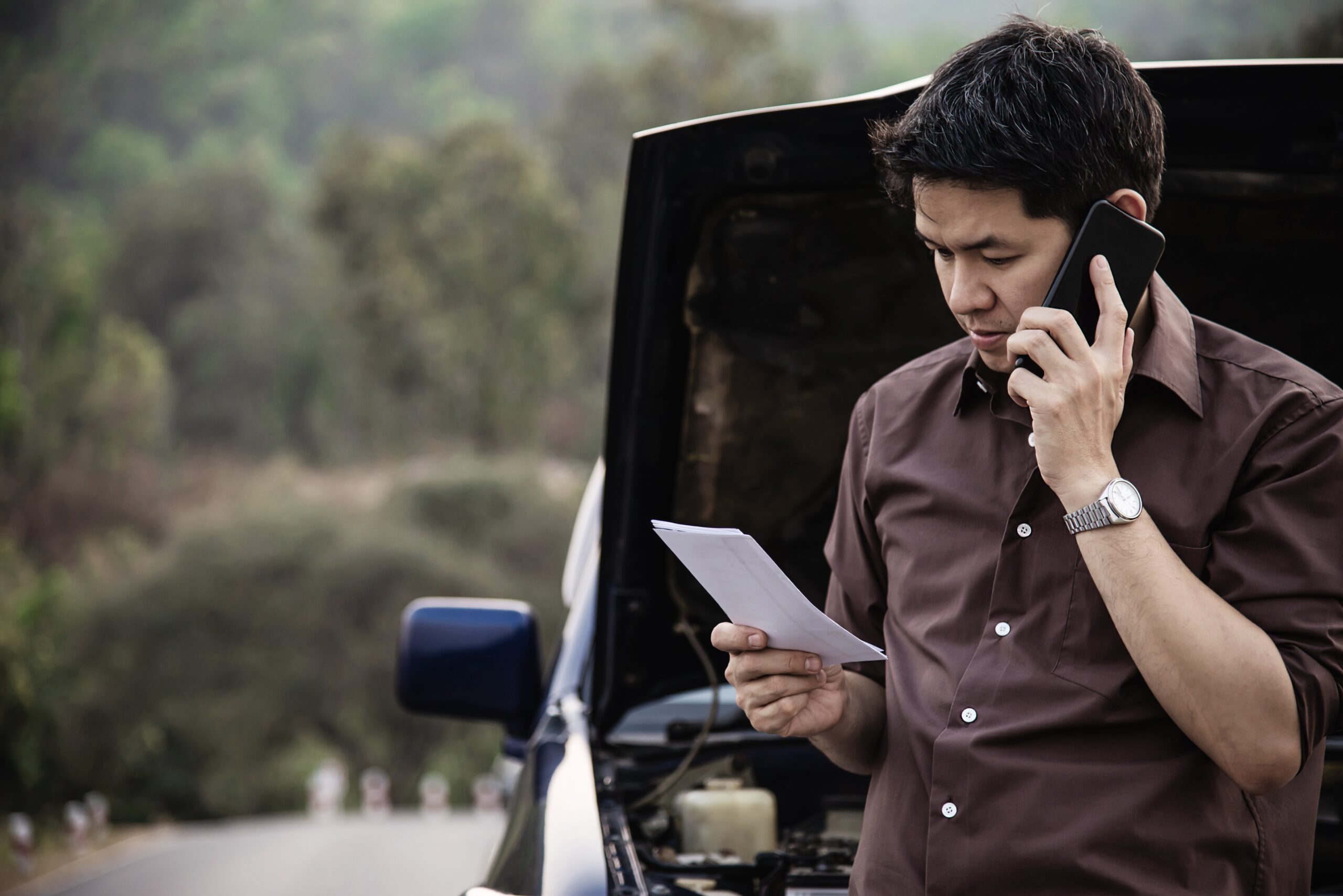 Man on the phone reviewing documents near a car with an open hood on a local road, illustrating the challenges of vehicle issues after a multi-car accident.