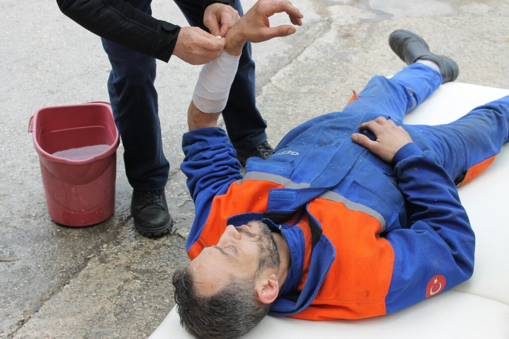 Injured worker receiving medical assistance on the ground, with a person applying a bandage to his arm, highlighting the importance of medical care after accidents.
