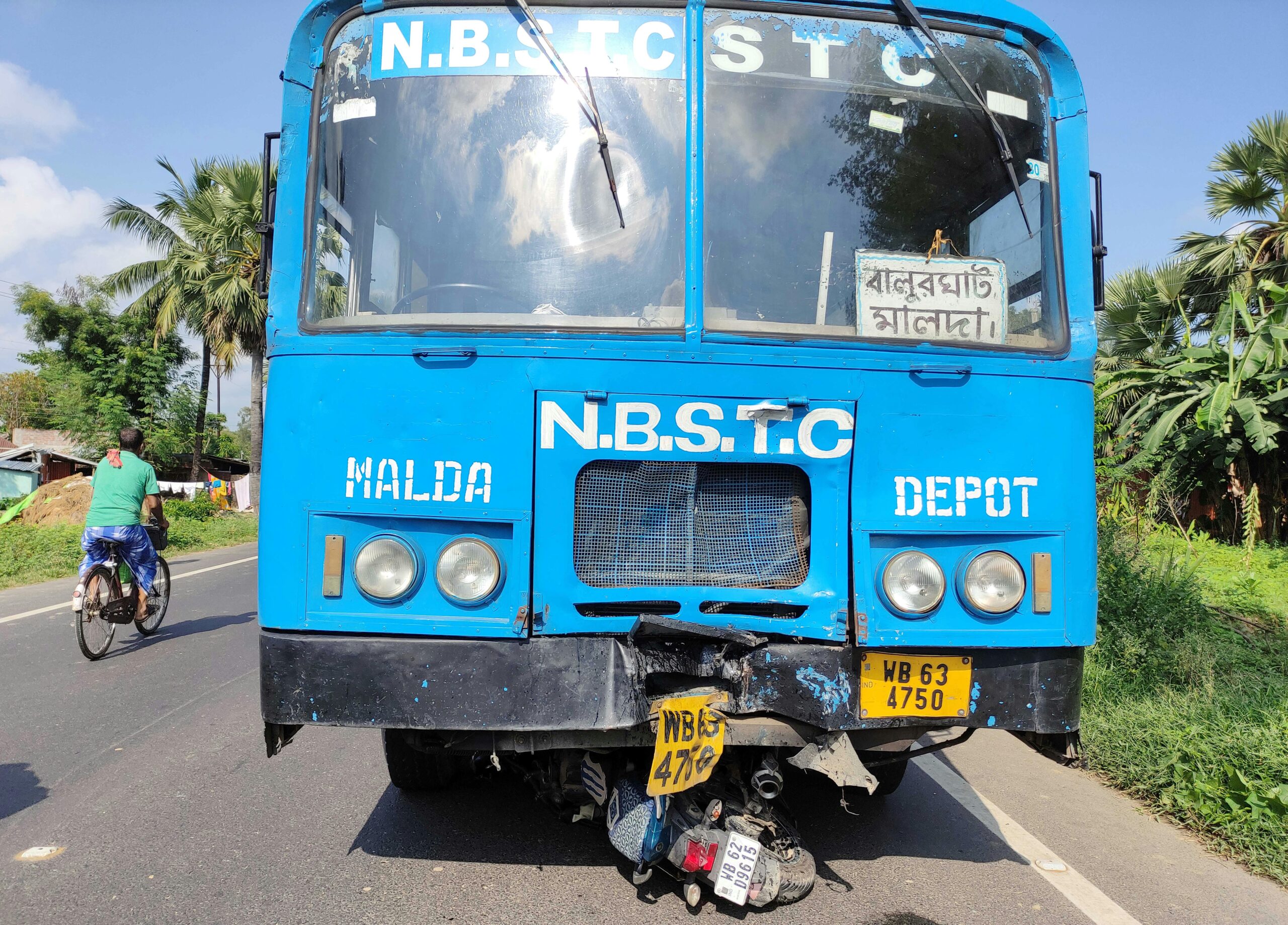 Blue bus with "N.B.S.T.C" and "MALDA" signage, showing damage to the front, parked on a rural road with a cyclist in the background, illustrating transportation and potential accident scenarios relevant to personal injury law in Wilson County.