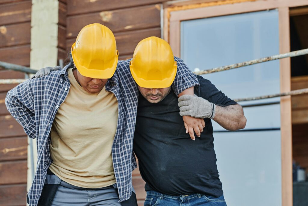 Construction workers assisting an injured colleague wearing yellow hard hats, highlighting workplace safety and personal injury scenarios relevant to legal claims in Little Forest Hills.
