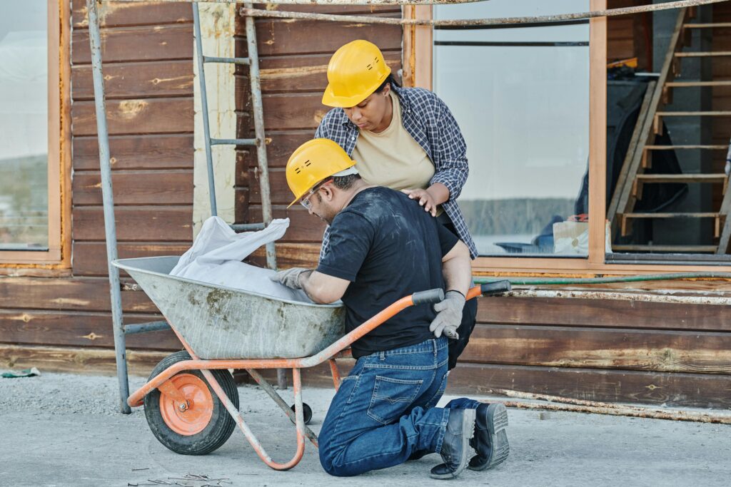 Two construction workers in hard hats, one kneeling beside a wheelbarrow filled with materials, while the other assists from behind, at a building site.