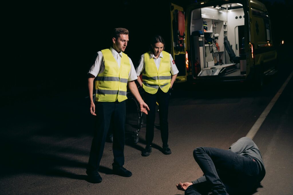 Emergency responders in reflective vests attending to a person lying on the road at night, with an ambulance in the background, illustrating the aftermath of a traffic accident.