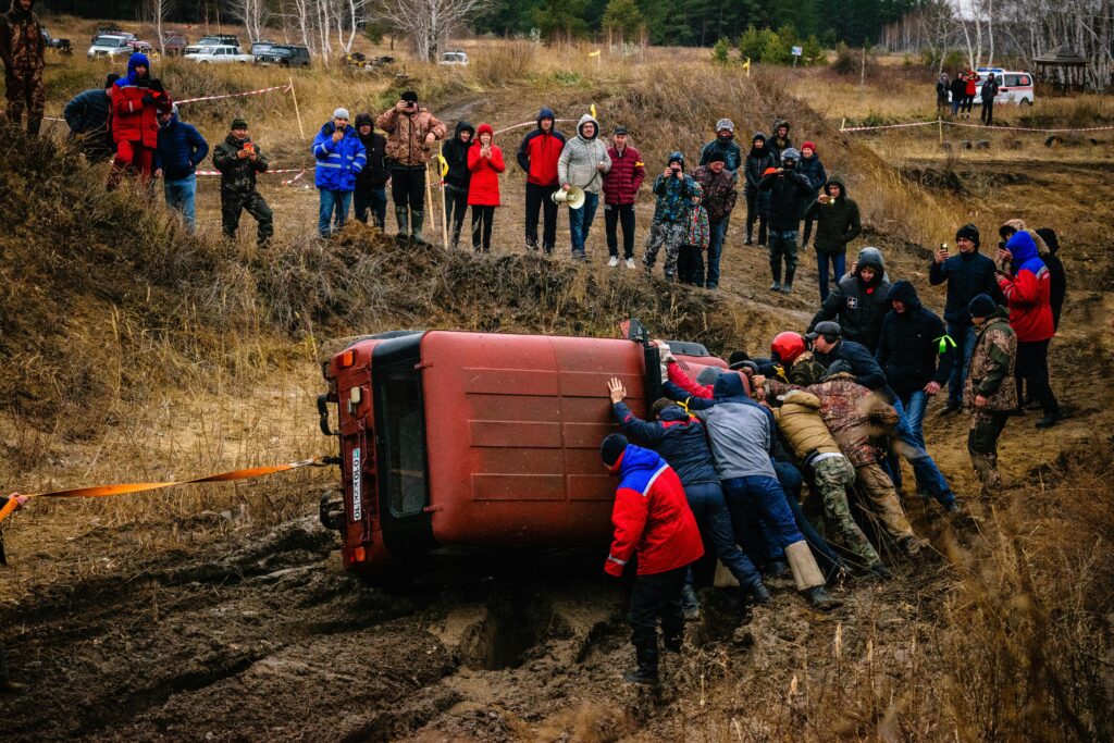 Group of people assisting in the recovery of a flipped vehicle in a muddy area, highlighting community support and teamwork in emergency situations.
