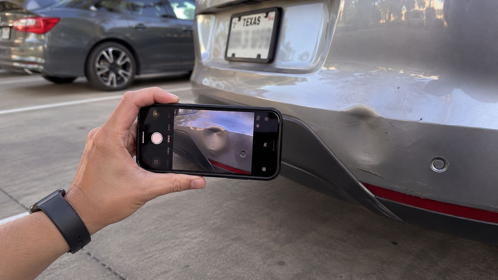 A person holding a smartphone to take a photo of a dented bumper on a silver car.
